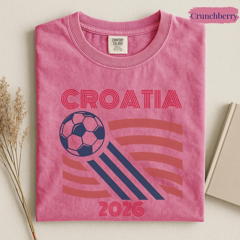 May include: A pink short-sleeved t-shirt featuring the text CROATIA in red, a dark blue soccer ball graphic with motion lines, and the year 2026 in red. The shirt has a Comfort Colors brand label on the inside of the collar and a pink tag.