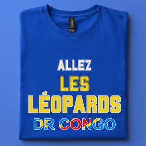 May include: A royal blue short-sleeved t-shirt featuring the white text ALLEZ, yellow text LES LÉOPARDS, and the text DR CONGO in blue, yellow, and red colors. The shirt has a crew neckline and a black brand label attached to the inner collar area.