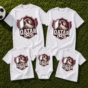 May include: White t-shirts and a onesie featuring a shield design with a falcon, soccer ball, and the text "QATAR 2026". The shirts are short-sleeved and suitable for a family. The shirts are on a green surface.