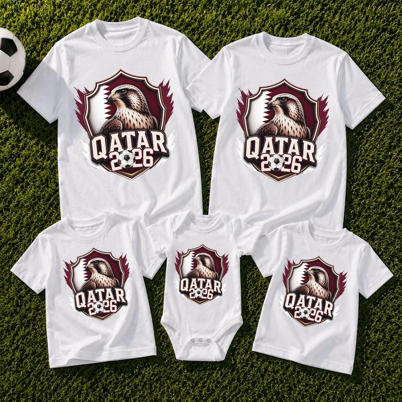 May include: White t-shirts and a onesie featuring a shield design with a falcon, soccer ball, and the text "QATAR 2026". The shirts are short-sleeved and suitable for a family. The shirts are on a green surface.
