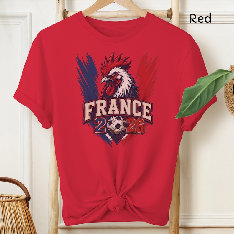 May include: Red t-shirt featuring a graphic of a rooster with the text "FRANCE 2026" and a soccer ball. The design incorporates the colors of the French flag: blue, white, and red. The shirt is displayed on a wooden hanger.