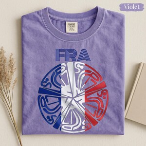 May include: A violet crew neck t-shirt featuring the text FRA in blue lines above a circular graphic. The graphic is divided into segments colored in blue, white, and red, resembling the French flag, with intricate white line art patterns.