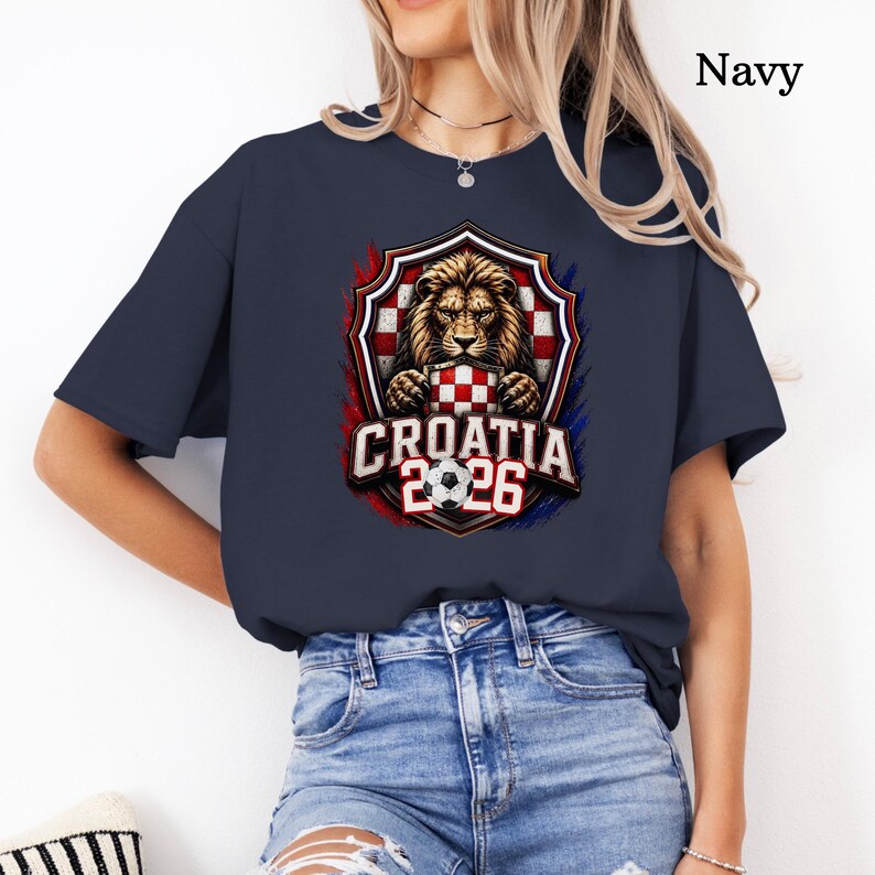 May include: Navy blue t-shirt featuring a shield design with a lion holding a checkered pattern, the Croatian flag. The text "CROATIA 2026" and a soccer ball are below the shield. The shirt is a crew neck.