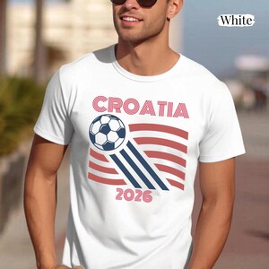 May include: A white short-sleeved t-shirt featuring a graphic design with the text CROATIA and 2026 in red. The design includes a black and white soccer ball with red and blue striped accents resembling a flag. The word White is printed in the corner.