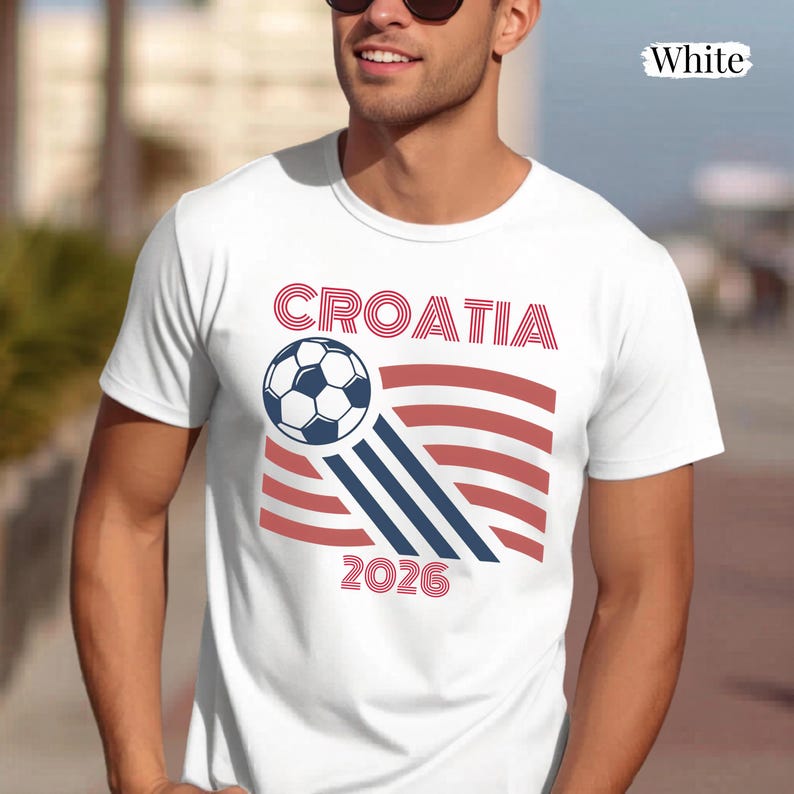May include: A white short-sleeved t-shirt featuring a graphic design with the text CROATIA and 2026 in red. The design includes a black and white soccer ball with red and blue striped accents resembling a flag. The word White is printed in the corner.