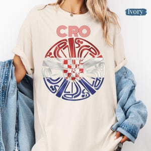 May include: An ivory t-shirt featuring a circular graphic with the text CRO at the top. The center contains a red, white, and blue design with a checkered shield pattern, surrounded by stylized, swirling lines in red, white, and blue colors.