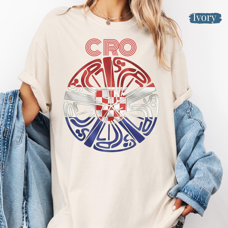 May include: An ivory t-shirt featuring a circular graphic with the text CRO at the top. The center contains a red, white, and blue design with a checkered shield pattern, surrounded by stylized, swirling lines in red, white, and blue colors.