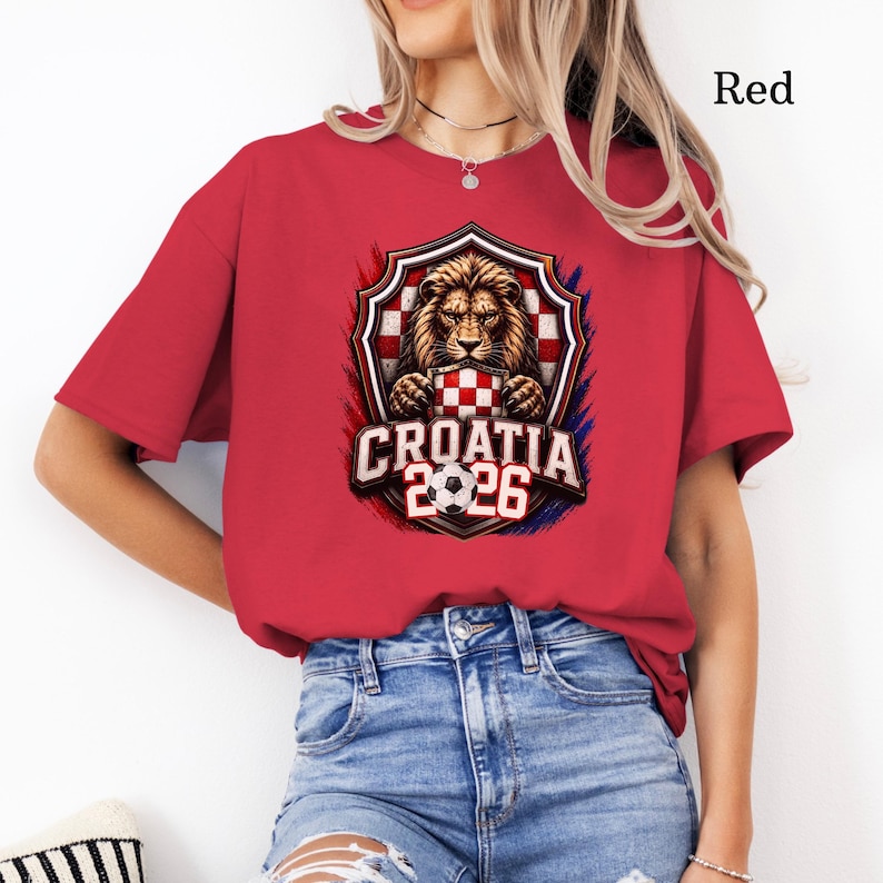 May include: Red t-shirt featuring a shield design with a lion, a checkered pattern, and the text "CROATIA 2026". The design is accented with red, white, and blue colors. The shirt is made of a soft material.