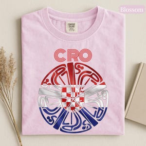 May include: A light pink t-shirt featuring the text CRO above a circular graphic. The graphic contains a red and white checkered pattern in the center, surrounded by stylized red, white, and blue lines forming a circular shape with the text CROATIA.