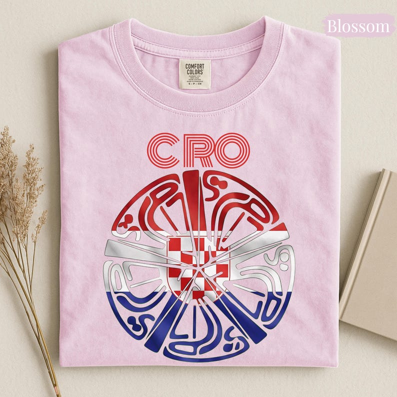 May include: A light pink t-shirt featuring the text CRO above a circular graphic. The graphic contains a red and white checkered pattern in the center, surrounded by stylized red, white, and blue lines forming a circular shape with the text CROATIA.