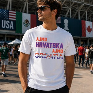 May include: A white short-sleeved t-shirt featuring the text AJMO in orange, HRVATSKA in purple, AJMO in orange, and CROATIA in blue and red checkered patterns. The shirt is worn by a person with dark hair and sunglasses in an outdoor setting.