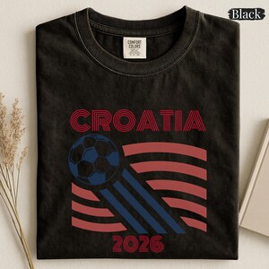 May include: A black crew neck t-shirt featuring the word CROATIA in red, a blue soccer ball graphic, and red wavy lines. The design includes three blue diagonal stripes and the year 2026 in red. The shirt has a Comfort Colors brand neck label.