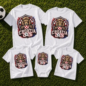 May include: White t-shirts and a onesie with a Croatia 2026 design. The design features a lion holding a soccer ball and a Croatian flag shield. The shirts are on a green grassy surface.