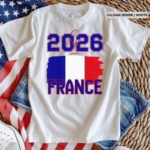 May include: White t-shirt with a blue and red design. The shirt features the number "2026" above a stylized French flag and the word "FRANCE" in blue and red. The shirt is made of white fabric.