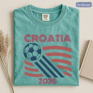 May include: A seafoam green t-shirt featuring the text CROATIA in red, a graphic of a soccer ball, three diagonal navy blue stripes, red curved lines, and the text 2026 in red. The shirt has a Comfort Colors brand label at the inner neckline.