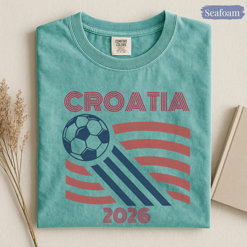 May include: A seafoam green t-shirt featuring the text CROATIA in red, a graphic of a soccer ball, three diagonal navy blue stripes, red curved lines, and the text 2026 in red. The shirt has a Comfort Colors brand label at the inner neckline.