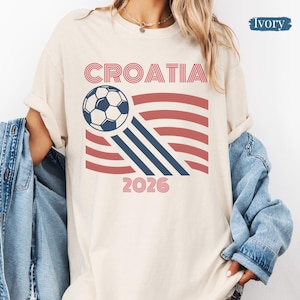 May include: An ivory short-sleeve t-shirt featuring a graphic print with the text CROATIA and 2026 in red. The design includes a soccer ball and red and blue curved lines. A blue denim jacket is draped over the shoulders of the t-shirt.