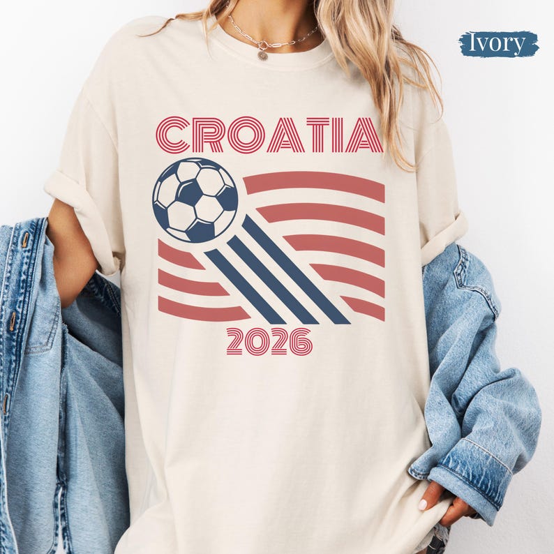 May include: An ivory short-sleeve t-shirt featuring a graphic print with the text CROATIA and 2026 in red. The design includes a soccer ball and red and blue curved lines. A blue denim jacket is draped over the shoulders of the t-shirt.