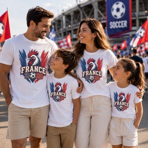 May include: A family of four wearing matching white t-shirts with a rooster graphic and the text "FRANCE 2026". The design incorporates the colors of the French flag and a soccer ball. The family is standing outside a stadium.