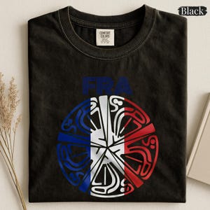 May include: A black crew neck t-shirt featuring the text "FRA" in blue above a circular graphic. The graphic is divided into vertical blue, white, and red stripes, overlaid with a stylized, intricate white line pattern resembling a Celtic knot.
