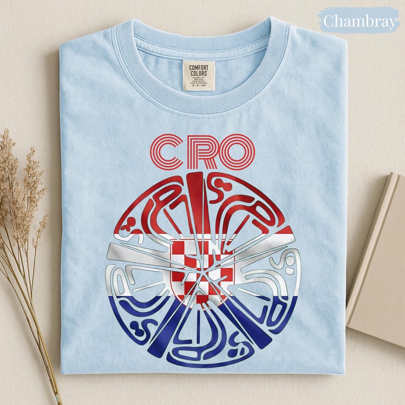 May include: A light blue crew neck t-shirt featuring the text CRO in red outlined letters above a circular graphic. The graphic contains a stylized red, white, and blue Croatian flag pattern with a red and white checkered shield in the center.