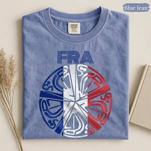 May include: A blue jean colored t-shirt featuring the text FRA in blue block letters above a circular graphic. The graphic displays a stylized design with blue, white, and red segments arranged in a radial pattern, resembling the French national flag.