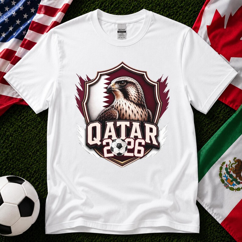 May include: White t-shirt featuring a shield design with a falcon illustration and the text "QATAR 2026" in a bold font. The design incorporates a soccer ball. Flags of the USA, Canada, and Mexico are in the background.