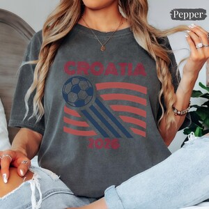 May include: A charcoal gray short-sleeve t-shirt featuring a graphic print with the text CROATIA and 2026. The design includes a dark blue soccer ball with motion lines and red horizontal stripes. The shirt has a crew neckline and a relaxed fit.