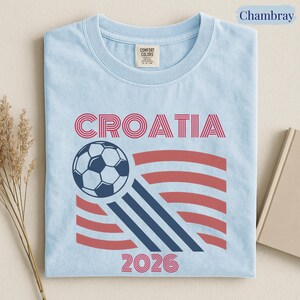 May include: A light blue t-shirt featuring the text CROATIA in red, a graphic of a soccer ball, and the year 2026 in red. The design includes red and dark blue striped accents. The shirt has a Comfort Colors brand label at the inner neckline.