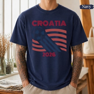 May include: A navy blue short-sleeve t-shirt featuring a graphic design with the text CROATIA and 2026. The design includes a stylized soccer ball and red and blue wavy lines. The shirt has a crew neckline and a standard fit.