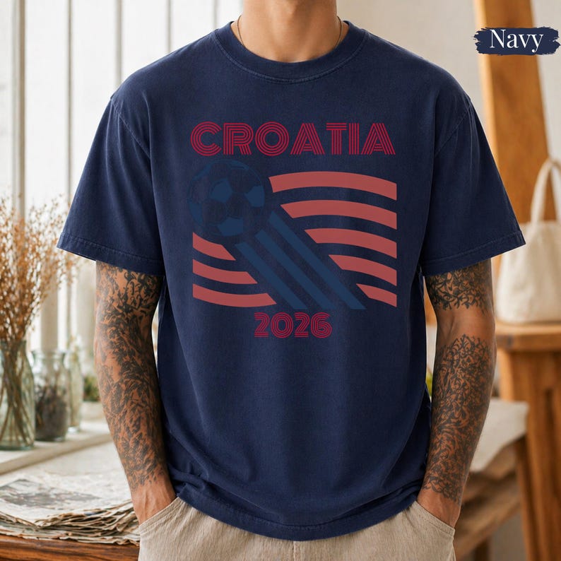 May include: A navy blue short-sleeve t-shirt featuring a graphic design with the text CROATIA and 2026. The design includes a stylized soccer ball and red and blue wavy lines. The shirt has a crew neckline and a standard fit.