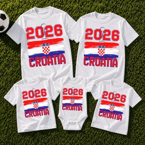 May include: White t-shirts and a onesie with a Croatian flag design. The shirts display the year "2026" in red, along with the word "CROATIA". The design includes red, white, and blue brushstrokes, and a checkered emblem.
