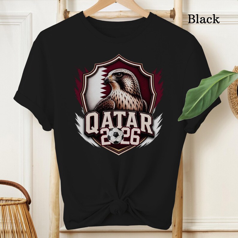 May include: Black t-shirt featuring a graphic of a falcon inside a shield with the Qatar flag and the text "QATAR 2026" and a soccer ball. The word "Black" is printed at the top right of the shirt.