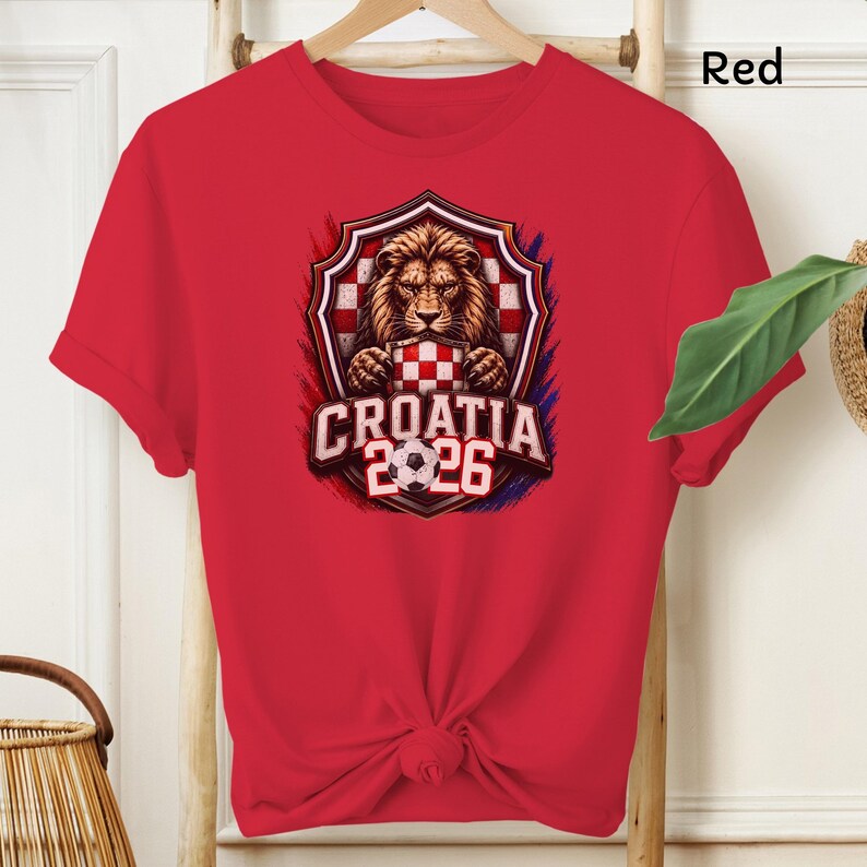 May include: Red t-shirt featuring a graphic of a lion holding a Croatian checkered shield and soccer ball. The text "CROATIA 2026" is below the shield. The shirt is hanging on a wooden hanger.