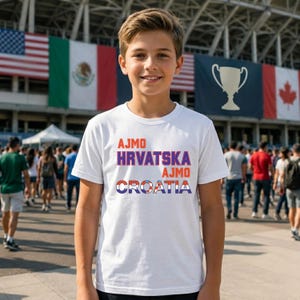 May include: A white short-sleeved t-shirt featuring the text AJMO HRVATSKA AJMO CROATIA printed on the front. The word AJMO is in orange, HRVATSKA is in purple, and the word CROATIA is stylized with the red and white Croatian flag pattern.