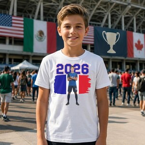 Może przedstawiać: Biała koszulka z grafiką piłkarza w niebieskim stroju, flagą Francji i numerem "2026" w kolorze niebieskim. Koszulka jest noszona przez młodą osobę przed stadionem z flagami.