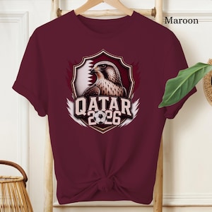 May include: Maroon t-shirt featuring a graphic of a falcon inside a shield with the Qatar flag and the text "QATAR 2026" and a soccer ball. The shirt is displayed on a wooden hanger.