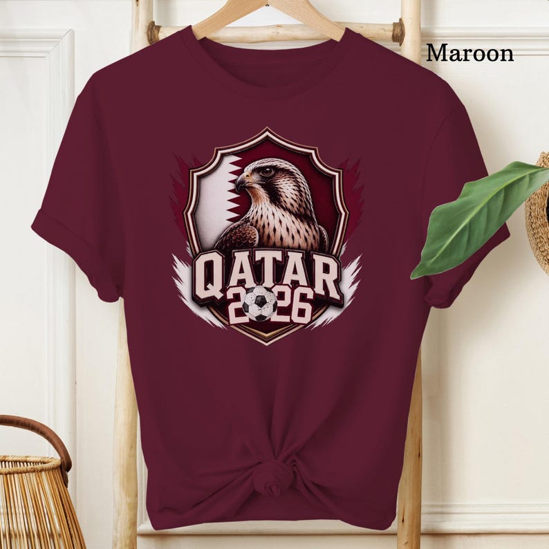 May include: Maroon t-shirt featuring a graphic of a falcon inside a shield with the Qatar flag and the text "QATAR 2026" and a soccer ball. The shirt is displayed on a wooden hanger.
