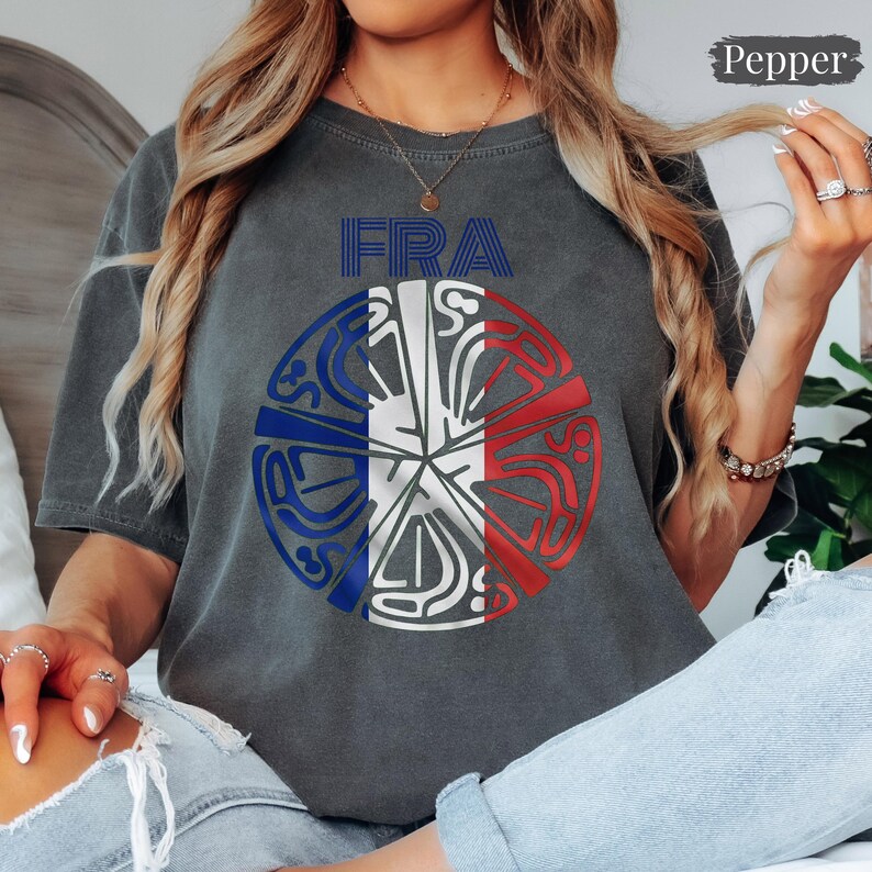 May include: A charcoal gray short-sleeve t-shirt featuring the text FRA in blue above a circular graphic. The graphic displays a stylized French flag design with blue, white, and red segments integrated with intricate, abstract line patterns.