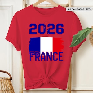 May include: A red t-shirt with the text "2026 FRANCE" in blue, featuring a stylized French flag design. The shirt is hanging on a wooden hanger.