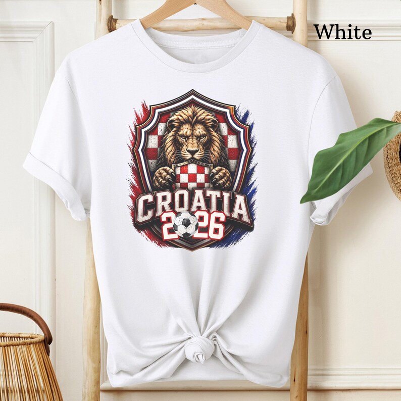 May include: White t-shirt featuring a graphic of a lion holding a checkered pattern, with the text "CROATIA 2026" and a soccer ball. The design is framed by red, white, and blue accents, evoking a national flag. The shirt is made of a soft material.
