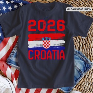 May include: Navy blue t-shirt with the text "2026 CROATIA" in red. The design includes a red, white, and blue stripe with the Croatian coat of arms. The shirt is a Gildan 5000B in navy.