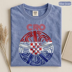 May include: A blue jean colored t-shirt with a crew neckline and a Comfort Colors label. The graphic features the red text CRO above a circular design containing the red, white, and blue Croatian flag pattern with stylized, swirling line art.