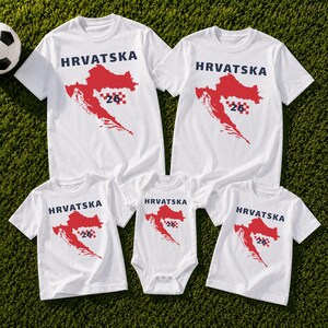 Op de afbeelding: Een collectie witte T-shirts met korte mouwen en &eacute;&eacute;n witte romper met korte mouwen, elk voorzien van een rode kaart van Kroati&euml;, de tekst HRVATSKA in donkerblauw en een klein rood-wit geblokt vierkant met het nummer 26 op de voorzijde.
