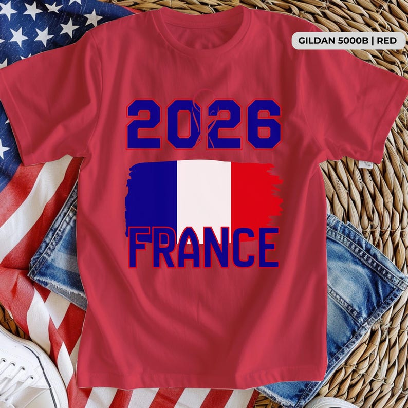 May include: Red Gildan t-shirt with a blue and white graphic of the French flag and the text "2026 FRANCE" in blue. The shirt is displayed on a background with an American flag and denim.