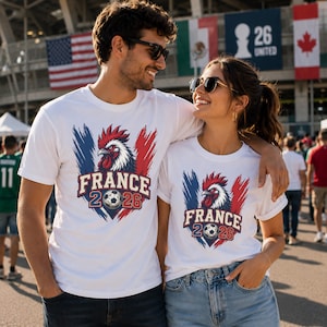 May include: Two white t-shirts with a graphic of a rooster in red, white, and blue, the colors of the French flag. The shirts also feature the text "FRANCE 2026" and a soccer ball. The shirts are worn by a man and a woman.