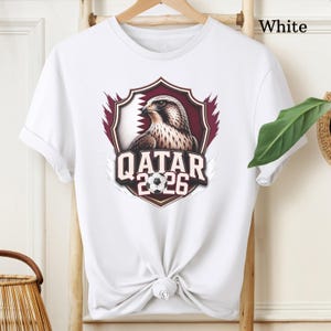 May include: White t-shirt featuring a graphic of a falcon inside a maroon shield with the Qatar flag and the text "QATAR 2026" and a soccer ball. The shirt is tied at the waist.