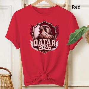 May include: Red t-shirt featuring a graphic of a falcon inside a shield with the text "QATAR 2026" and a soccer ball. The shirt is made of a soft material and has short sleeves.