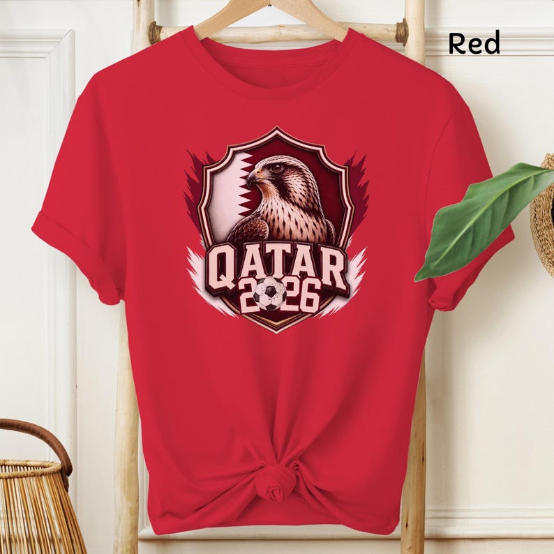 May include: Red t-shirt featuring a graphic of a falcon inside a shield with the text "QATAR 2026" and a soccer ball. The shirt is made of a soft material and has short sleeves.
