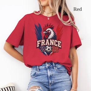 May include: Red t-shirt featuring a graphic of a rooster with the text "FRANCE 2026" and a soccer ball. The design incorporates blue and red brushstrokes, representing the French flag. The shirt is a solid red color.